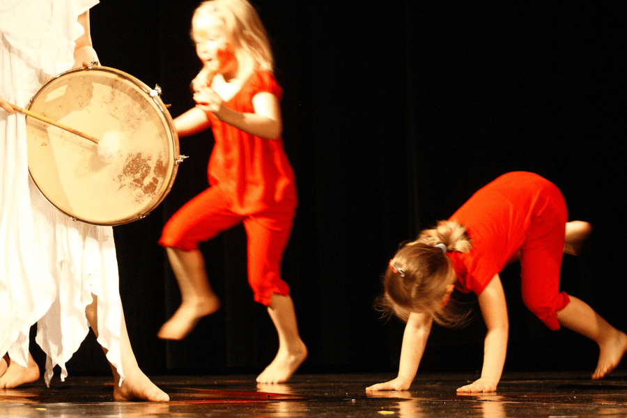 Kinder in roten Kostümen führen ein Tanzstück auf, während ein Kind in einem weißen Kostüm die Trommel schlägt.