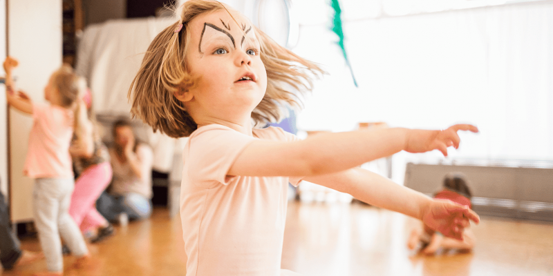 Ein Kind etwa 4, 5Jahre alt, tanzt im Tanzraum und wirft eine grüne Feder hoch. Im Hintergrund sieht man weitere Kinder und die Tanzlehrerin