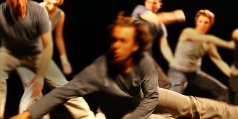 Gruppe von Tänzer*innen, im Vordergrund gleitet ein junger Mann dynamisch zum Boden, im Hintergrund weiter Tänzerinnen und Tänzer die wohl folgen