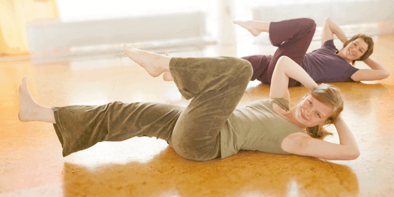 zwei Frauen üben in Rückenlage die Pilatesübung criss cross