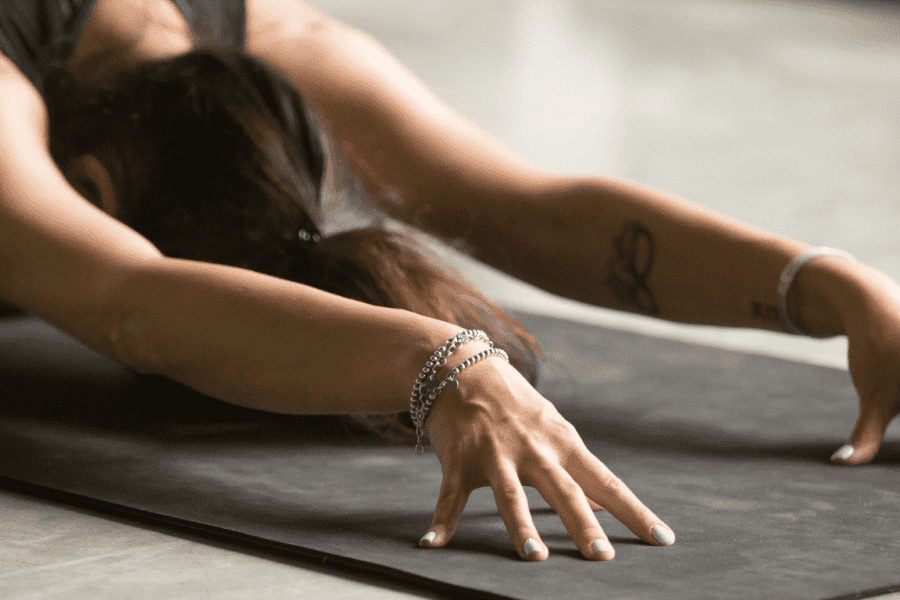 Eine Frau übt eine Rückendehnung auf einer Pilatesmatte. Sie sitzt dazu auf ihren Fersen und streckt Oberkörper und Arme nach vorne-unten aus. Ihre Fingerspitzen sind aufgestellt.