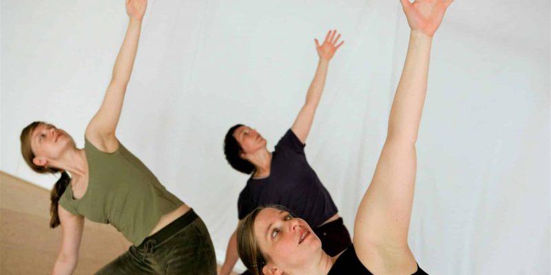 drei Yogaübende stehen in Trikonasana, einer Yogastehhaltung. Der Blick nach oben gerichtet und ein Arm nach oben gestreckt.
