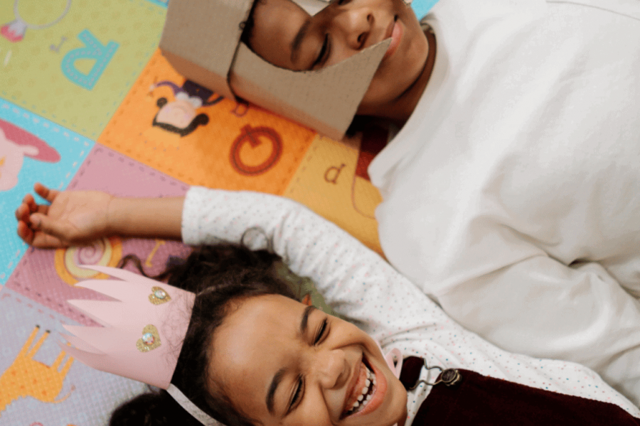 Ein Mädchen mit einer selbst gebastelten rosa Krone und ein Junge mit einem selbst gebastelten Ritterhelm liegen lachend auf einem bunten Spielteppich.
