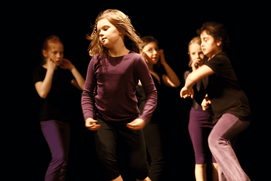 Ein Mädchen im Alter von 9 Jahren ist auf einer Bühne zu sehen, wie sie tanzt. Sie ist mitten im Schritt, die Hände zu einer Geste, als wollten alle Fingerspitzen zueinander geführt werden. Im Hintergrund sind weitere Mädchen zu sehen, alle schwarz-lila angezogen.